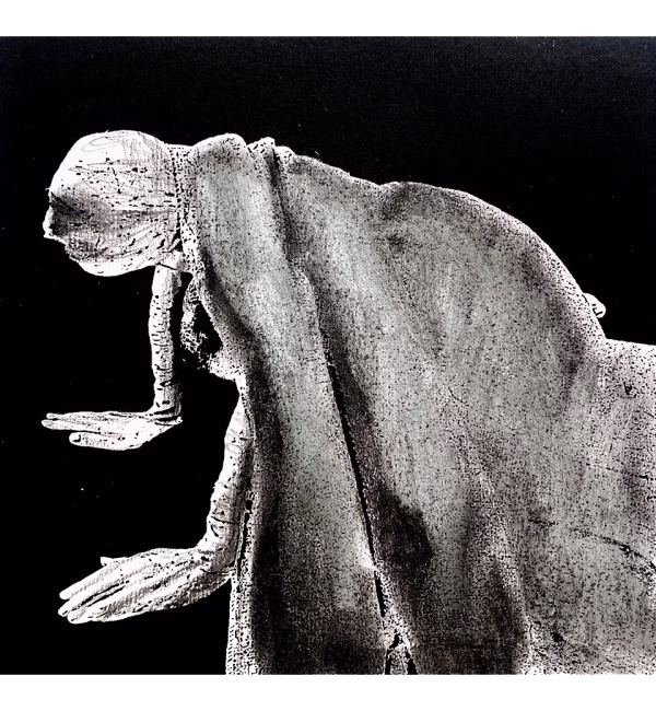 A woman crawls along the ground. She is bald, skeletal and covered in a removal blanket. Printed and tinted/shaded in black.