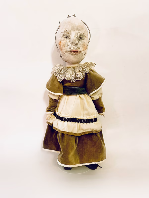 A doll wearing a sage green velvet and cream cotton dress. With a kilner jar clip over its head.
