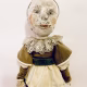 A doll wearing a sage green velvet and cream cotton dress. With a kilner jar clip over its head.