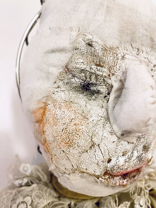 A doll wearing a sage green velvet and cream cotton dress. With a kilner jar clip over its head. Head view close-up.