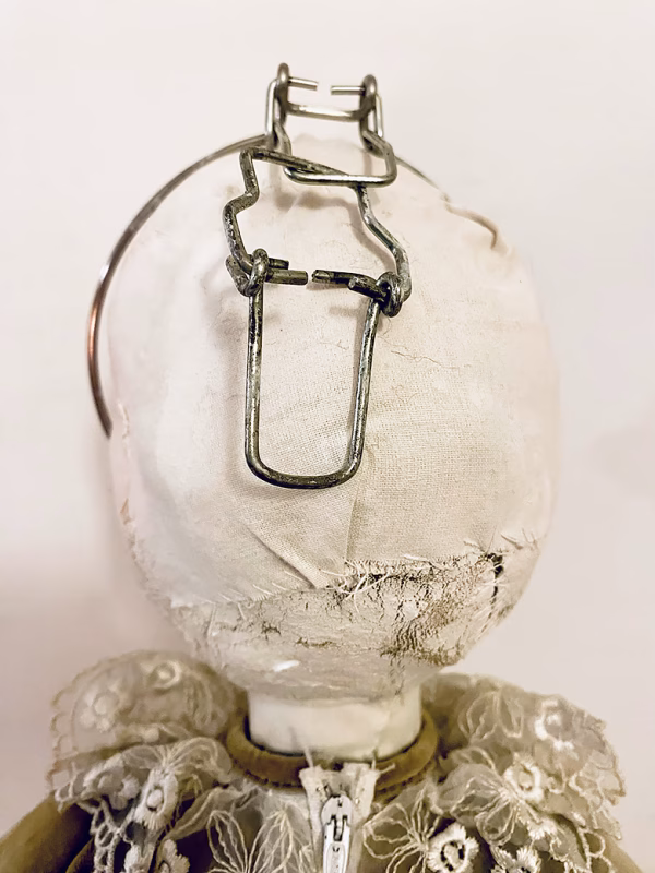 A doll wearing a sage green velvet and cream cotton dress. With a kilner jar clip over its head. Head view close-up.