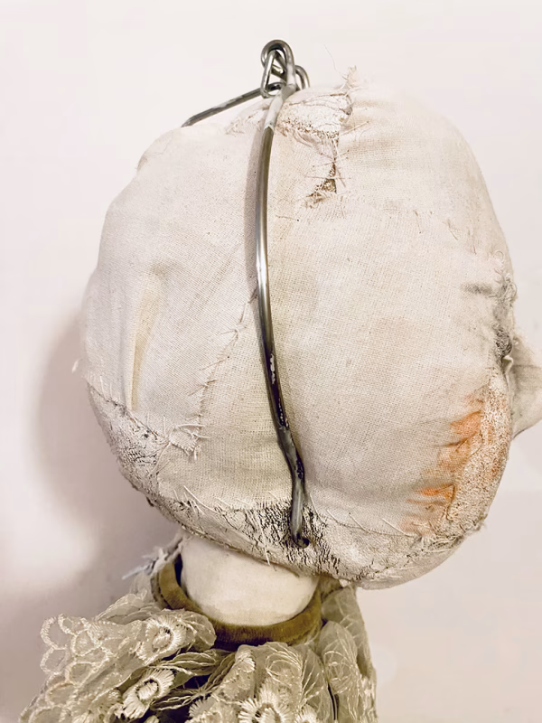 A doll wearing a sage green velvet and cream cotton dress. With a kilner jar clip over its head. Head view close-up.