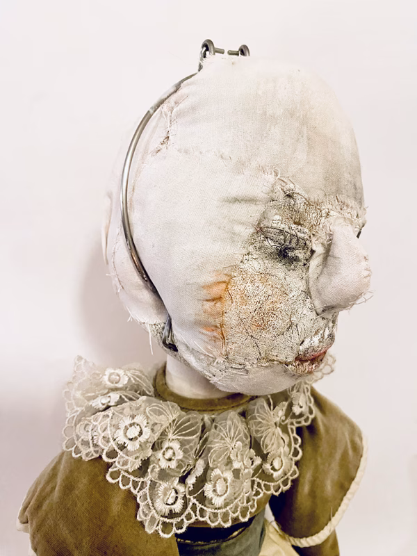 A doll wearing a sage green velvet and cream cotton dress. With a kilner jar clip over its head.