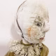 A doll wearing a sage green velvet and cream cotton dress. With a kilner jar clip over its head.