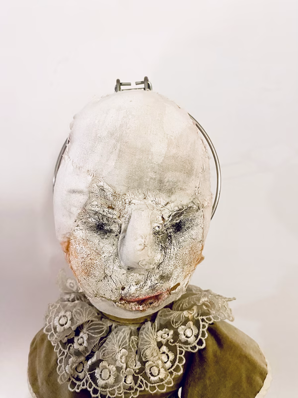 A doll wearing a sage green velvet and cream cotton dress. With a kilner jar clip over its head.