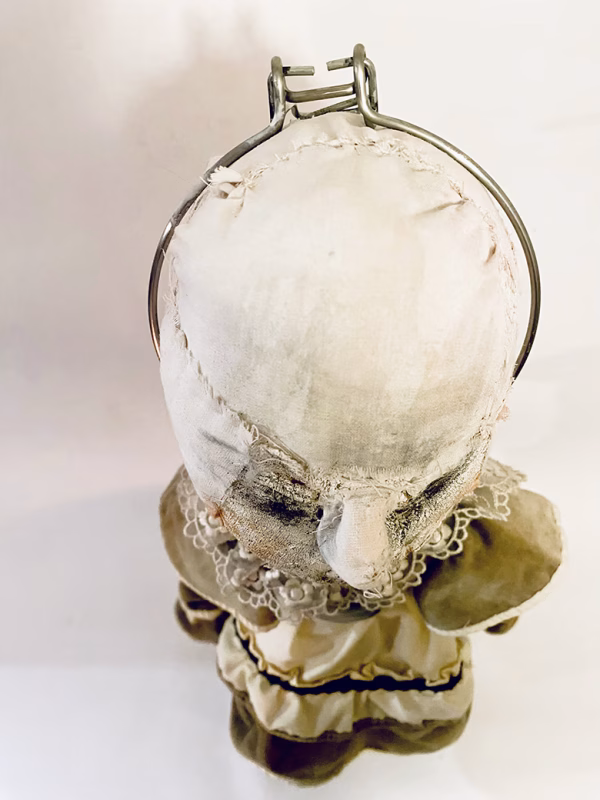 A doll wearing a sage green velvet and cream cotton dress. With a kilner jar clip over its head.